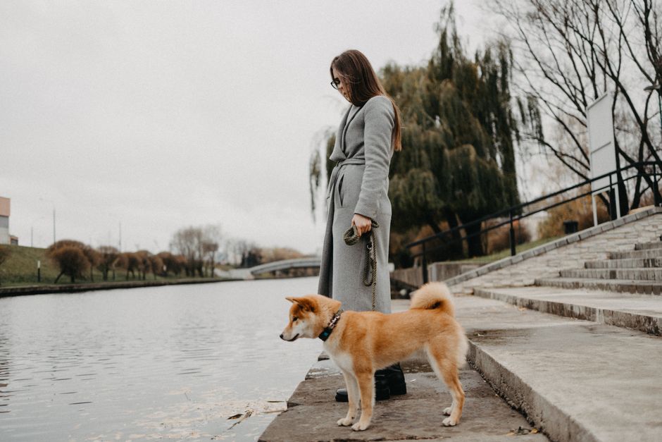 A woman dressed in a long grey coat is standing on a paved riverside walkway at Strand on the Green, holding a leash attached to a small, fluffy, reddish-brown dog with a white chest and paws. The wom