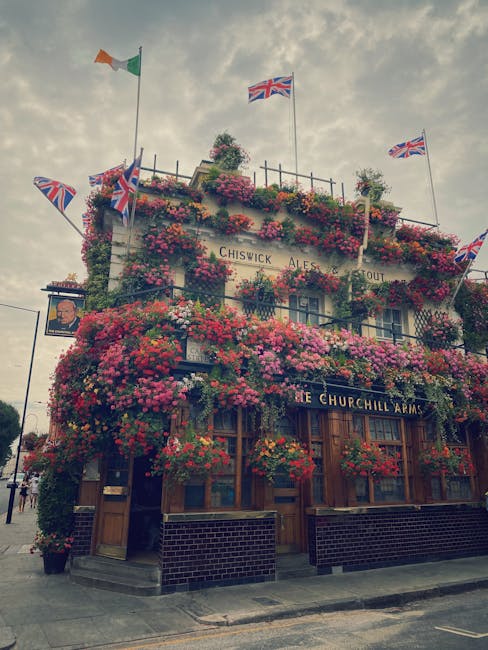 The exterior of the Churchill Arms pub in Chiswick is covered in vibrant, overflowing floral displays featuring pink, red, and purple blossoms, cascading from window boxes and hanging baskets. Union J
